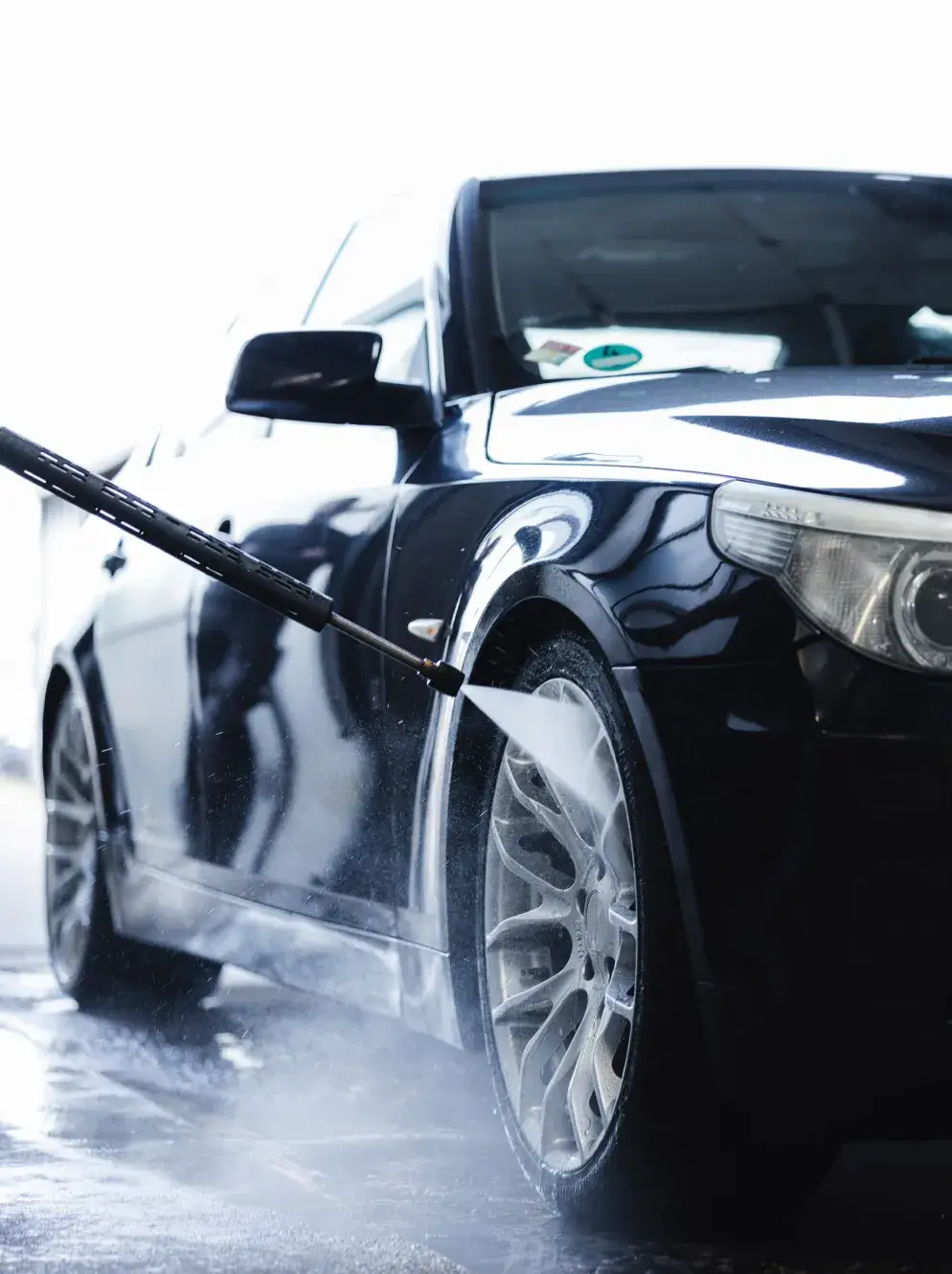 Ein dunkler Pkw wird mit einem Hochdruckreiniger gründlich an Reifen und Karosserie gewaschen - Fahrzeugpflege bei Karosserie Blanke.